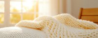 Cozy knitted cream blanket on bed near window with warm sunlight streaming through, creating peaceful morning atmosphere for lifestyle and home decor concepts.