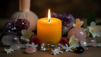 Burning orange candle surrounded by healing crystals, gemstones and white flowers creating peaceful spa atmosphere for meditation and wellness rituals.