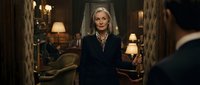 Confident blonde businesswoman in elegant black suit standing in luxurious hotel lobby with warm lighting and classic interior design.