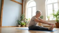 Senior woman with gray hair and light skin tone performing forward fold stretch on yoga mat in bright, plant-filled studio space. Wellness and fitness content.