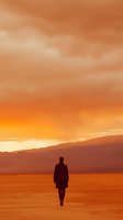 Silhouette of person walking alone across desert landscape at dramatic orange sunset with mountain range in background and moody cloudy sky.