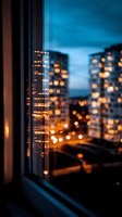Blurred city lights through window at dusk create dreamy bokeh effect with warm orange glow against blue hour sky, urban nightscape view from apartment.