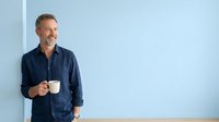 Middle-aged Caucasian man in casual navy blue shirt holding white coffee mug against light blue wall background, suitable for lifestyle and business content.