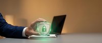 Businessman in dark suit holding glowing green security shield with padlock icon over laptop, representing data protection and cybersecurity solutions for business.