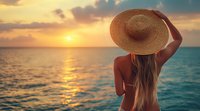 Young woman in straw hat watching golden sunset over calm ocean waters, long blonde hair flowing in breeze, back view creates serene summer vacation mood.