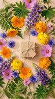 Kraft gift box tied with twine surrounded by colorful spring flowers including ranunculus, lupine, cornflower, and cosmos on sandy background.