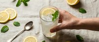 Hand holding refreshing lemon mint drink with ice cubes on rustic wooden table. Summer beverage with fresh citrus slices and green herbs for healthy lifestyle.