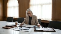 Senior professional woman with white hair and glasses, wearing beige blazer, reviewing documents at marble desk in modern office with large windows and contemporary furniture.