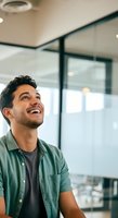 Young man with dark hair and beard, South Asian ethnicity, wearing casual green shirt over dark t-shirt, smiling while looking upward in modern office space with glass partitions and natural lighting.