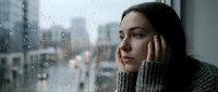 Young woman in cozy sweater looking thoughtfully through rain-covered window on gloomy day, perfect for mental health and wellness content.