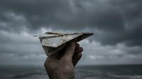 Hand holding paper airplane against dramatic stormy sky with dark clouds, symbolizing hope, dreams, and perseverance through challenges.