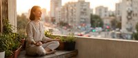 Woman with reddish-blonde hair sitting cross-legged on urban balcony ledge at sunset, overlooking cityscape with potted plants, contemplative pose for wellness and lifestyle content.