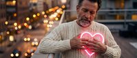Middle-aged Caucasian man in beige sweater holding glowing pink neon heart symbol with warm bokeh lights background for love and romance concepts.