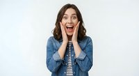Excited young woman with hands on cheeks showing surprise and joy, wearing denim jacket over striped shirt on white background for marketing campaigns.