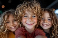 Three cheerful children with curly blonde hair and bright smiles looking at camera. Close-up portrait showing genuine happiness and friendship in natural lighting.
