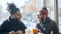 Two young African American men with beards and glasses sharing genuine laughter over coffee at cafe window. Natural daylight creates warm atmosphere for friendship moment.