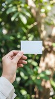 Hand holding a blank white card outdoors with lush green bokeh background, mockup for business card, invitation or message design.