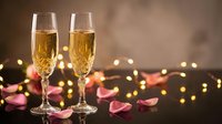 Two champagne flutes filled with golden bubbly surrounded by rose petals and warm bokeh lights creating romantic atmosphere for celebration or anniversary.