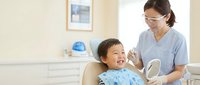 Young Asian boy smiling during dental checkup with female dentist in protective mask at modern clinic office for pediatric oral health care.