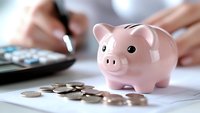 Pink ceramic piggy bank with scattered coins on financial documents while person calculates budget in background, representing savings and money management.