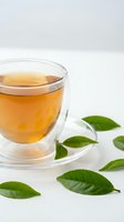 Clear glass teacup with golden herbal tea surrounded by fresh green tea leaves on white background for wellness and relaxation concepts.
