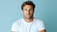 Portrait of athletic Caucasian male model with stubble beard wearing white t-shirt against blue background, looking directly at camera with confident expression.