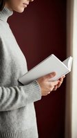 Young woman in cozy gray sweater reading open book by window with warm natural light creating peaceful atmosphere for literature and learning.