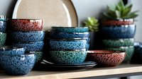 Colorful ceramic bowls with speckled glaze finish stacked on wooden shelf. Handmade pottery dinnerware collection in blue, green, brown tones for kitchen decor.