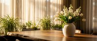 Sunlit wooden table with white lily of the valley flowers in ceramic vase and green plants by window with warm natural light streaming through curtains.