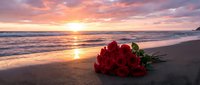 Red roses bouquet on sandy beach at sunset with golden sky reflection on ocean waves creating romantic atmosphere for love and Valentine's Day celebrations.