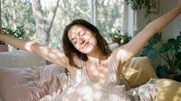 Young woman with medium-length dark hair and light skin tone stretching in bed near window with plants, wearing light pajamas, morning wellness and relaxation concept.