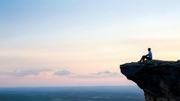 Silhouette of person sitting on cliff edge overlooking vast landscape during sunset, peaceful contemplative moment against pastel sky and distant horizon.