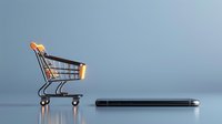 Empty miniature shopping cart next to smartphone on reflective surface against blue background, highlighting online shopping and e-commerce concept.