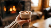 Cozy winter morning with steaming hot chocolate topped with marshmallows held in hands wearing knit sweater by warm fireplace in rustic living room.