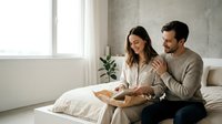 Happy couple enjoying breakfast in bed together in modern minimalist bedroom with natural light streaming through windows creating intimate morning moment.