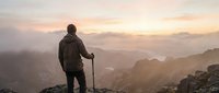 Hiker with trekking pole standing on mountain peak at sunrise, overlooking misty valley landscape below for adventure and outdoor recreation content.