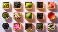 Various tea types and ingredients in wooden square containers on pink background, including matcha powder, loose leaf tea, dried flowers and nuts arranged in geometric pattern.
