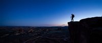 Silhouette of hiker standing on rocky cliff edge during twilight hour with desert landscape below and gradient sky transitioning from deep blue to purple.