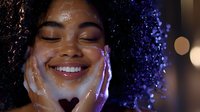 Young African American woman with curly hair smiling with eyes closed while holding face in hands, illuminated by purple and blue neon lights creating magical atmosphere.