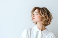 Young woman with wavy blonde bob hairstyle wearing white blouse against light background, profile view showing natural makeup and modern casual style.