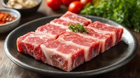 Fresh raw beef short ribs with marbling on dark plate ready for grilling or braising with herbs and vegetables in background for cooking preparation.