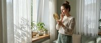 Woman with brown hair in casual beige shirt and green pants holding mirror near sunny window with potted plants and sheer curtains, morning skincare routine.