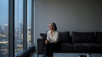 Professional businesswoman sitting on modern sofa in luxury office with city skyline view through floor-to-ceiling windows during evening hours.