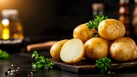 Fresh yellow potatoes with cut half showing white flesh, garnished with parsley and peppercorns on rustic wooden board against dark background. Food photography.