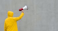 Person in bright yellow hoodie holding white megaphone against concrete wall for communication and announcement concepts.