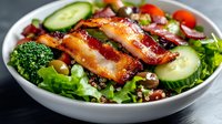 Fresh mixed greens salad topped with crispy bacon strips, cucumber slices, cherry tomatoes and broccoli florets in white bowl for healthy dining concept.