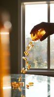 Hand pouring golden vitamin capsules from amber bottle onto surface near bright window with natural light and blurred background.