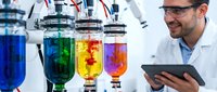 Male scientist in white lab coat and safety glasses examining colorful chemical solutions in laboratory glassware while using digital tablet for research analysis.