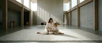 Young Asian woman practicing meditation in minimalist modern interior with natural light streaming through large windows onto woven mat.
