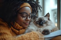 Young African American woman in glasses and orange beanie relaxing near window with Birman cat. Cozy home atmosphere with natural lighting and warm autumn sweater.